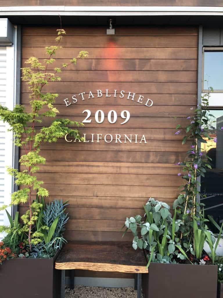 A rustic wooden wall displays the text "Established 2009 California" in white. Surrounding plants, including green foliage and succulents, frame the bench at the base of the wall, adding a lush, natural touch to the setting. CA Norml