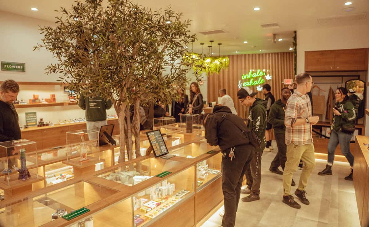 Customers browse products in a modern cannabis dispensary with wooden display cases and a tree in the center. A neon sign on the wall reads "inhale/exhale." People are engaged, looking at items, and interacting with staff. The atmosphere at Green Earth Collective appears casual and inviting. CA Norml