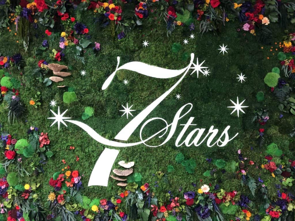 A lush green backdrop adorned with colorful flowers and greenery displays an elegantly designed white "7 Stars" logo. Sparkling star accents surround the number seven, adding a magical touch to the vibrant setting. CA Norml