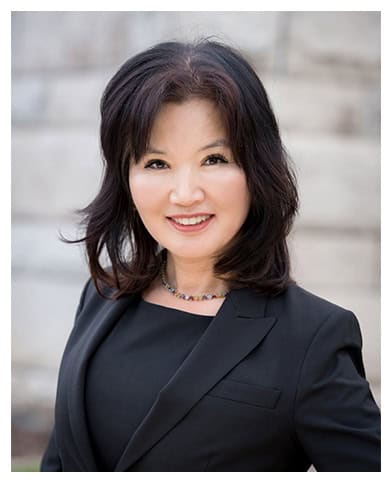 A woman with shoulder-length dark hair is smiling at the camera. She is wearing a black blazer and a beaded necklace. The background, a blurred, light-colored wall, hints at the professional setting of the Law Offices of Glew & Kim. CA Norml