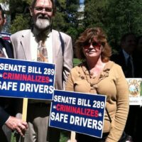 Two people holding signs protesting Senate Bill 289, which they claim criminalizes safe drivers. The man on the left is wearing a beige suit and tie, and the woman on the right is wearing sunglasses and a beige sweater. Trees and others are visible in the background of this 2013 Cal NORML news archive photo. CA Norml