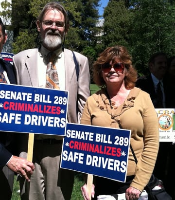 Two people holding signs protesting Senate Bill 289, which they claim criminalizes safe drivers. The man on the left is wearing a beige suit and tie, and the woman on the right is wearing sunglasses and a beige sweater. Trees and others are visible in the background of this 2013 Cal NORML news archive photo. CA Norml