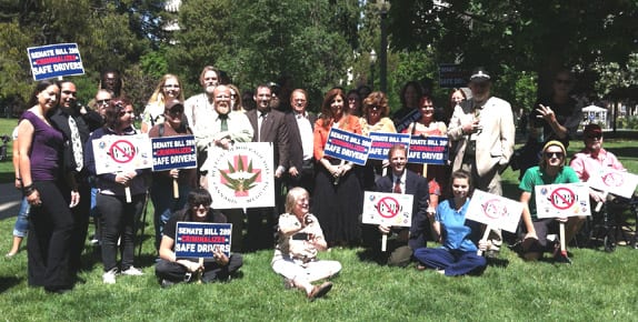 A group of Californians is gathered outside on a sunny day, holding signs and banners supporting Senate Bill 289, the Zero-Tolerance DUI Bill promoting safe driving. The diverse group includes people of various ages, all smiling and posing for the photo on a grassy area. CA Norml