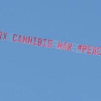 A red aerial banner towed by an airplane against a clear blue sky reads, "Holder: End RX Cannabis War #Peace4Patients," as medical marijuana protesters cheer below. CA Norml