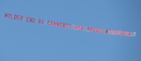 A red aerial banner towed by an airplane against a clear blue sky reads, "Holder: End RX Cannabis War #Peace4Patients," as medical marijuana protesters cheer below. CA Norml