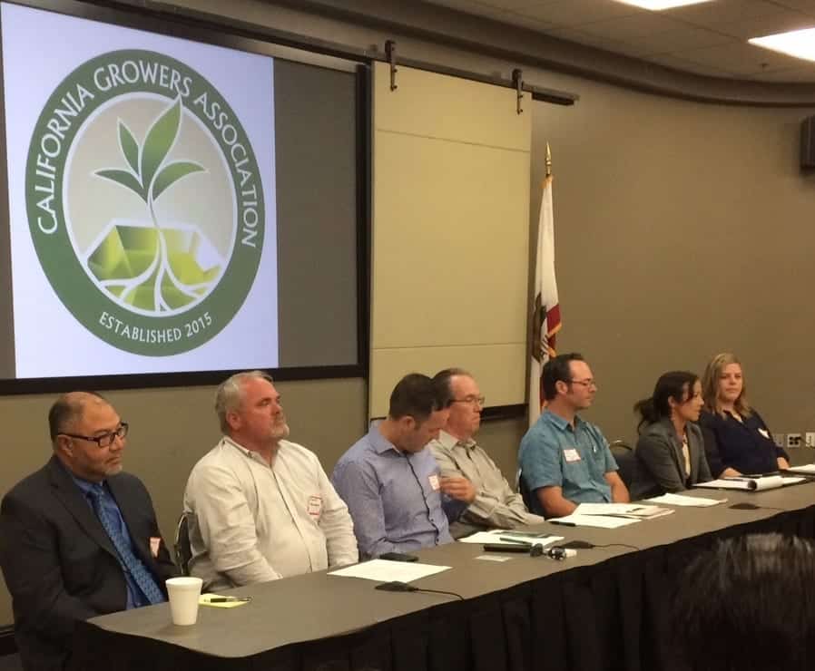 Seven people sit at a long table in a conference room, facing an audience. Behind them, the screen displays the "California Growers Association" logo. The individuals, including cannabis farmers and regulators, appear to be participating in a panel discussion. An American flag is visible in the background. CA Norml