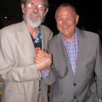 Two elderly men standing close together and smiling at the camera. They are shaking hands. Both are wearing suits; the man on the left has a beard and glasses and is wearing a light-colored suit, while the man on the right, resembling one of California's Senators, is in a checkered shirt and gray suit. CA Norml