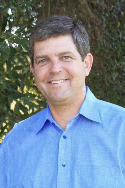 A smiling man with short dark hair is standing outdoors against a backdrop of greenery. Wearing a light blue button-down shirt, he is illuminated by sunlight that brightens his face and the surrounding leaves. This image, part of the 2015 News Archive, captures him in a candid moment. CA Norml