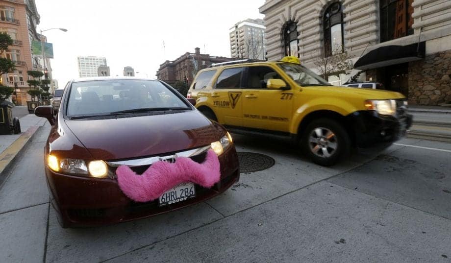 A red car with a large pink mustache on its front bumper is parked on the street in SF. A yellow taxi is driving past it on the right-hand side. The buildings and street in the background indicate an urban setting. CA Norml