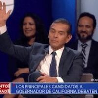 A man in a suit and tie raises his right hand during a debate among California gubernatorial candidates. He is seated on a stage with other individuals in the background. The text at the bottom reads "Los Principales Candidatos a Gobernador de California Debaten" and a logo for Univision is visible. CA Norml