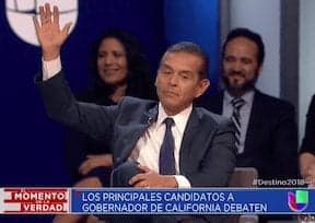 A man in a suit and tie raises his right hand during a debate among California gubernatorial candidates. He is seated on a stage with other individuals in the background. The text at the bottom reads "Los Principales Candidatos a Gobernador de California Debaten" and a logo for Univision is visible. CA Norml