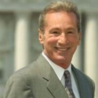 A smiling man with short, light-colored hair, wearing a light gray suit, white shirt, and a patterned tie, stands outdoors in front of an out-of-focus background. He appears confident and ready to discuss the latest developments in the Assembly regarding the Ammiano Medical Marijuana Regulation Bill. CA Norml