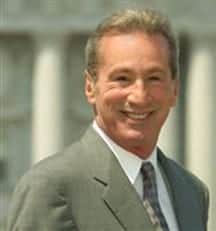 A smiling man with short, light-colored hair, wearing a light gray suit, white shirt, and a patterned tie, stands outdoors in front of an out-of-focus background. He appears confident and ready to discuss the latest developments in the Assembly regarding the Ammiano Medical Marijuana Regulation Bill. CA Norml