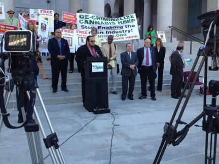 A group of people stand on the steps of a building with columns, holding signs advocating for marijuana decriminalization in California. A man speaks at a podium with cameras set up in the foreground to record the event. The signs include messages like "DE-CRIMINALIZE CANNABIS HEMP. CA Norml