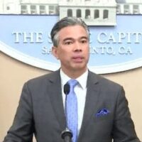 A man in a suit speaks at a podium with microphones in front of him. Behind him is a circular emblem with text stating "THE CAPITOL SACRAMENTO, CA." American flags are visible on either side of him as he discusses California's marijuana reform from 2018. CA Norml