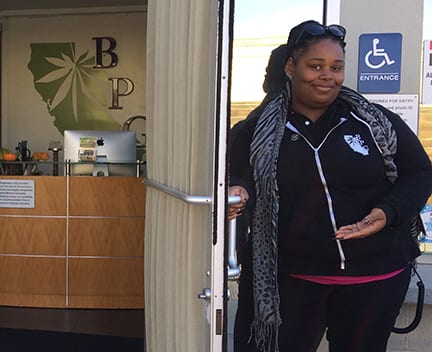 A woman stands in a doorway, holding the door open with one hand and gesturing inside with the other. She is wearing a black jacket with white details, a patterned scarf, and sunglasses on her head. Behind her, a reception desk and a sign with a leaf design promote California's recreational marijuana sales starting January 1. CA Norml