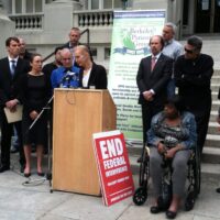 A group of men and women stand together on the steps of a building. One woman, speaking at a podium with microphones, has Cal NORML supporters standing behind her. A sign in front reads "END FEDERAL INTERFERENCE." The backdrop includes banners from the Berkeley Patients Group Medical Cannabis Dispensary. CA Norml