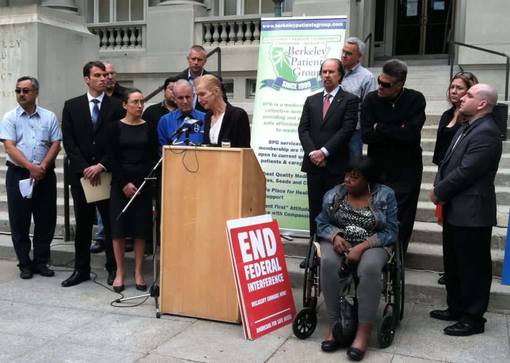 A group of men and women stand together on the steps of a building. One woman, speaking at a podium with microphones, has Cal NORML supporters standing behind her. A sign in front reads "END FEDERAL INTERFERENCE." The backdrop includes banners from the Berkeley Patients Group Medical Cannabis Dispensary. CA Norml