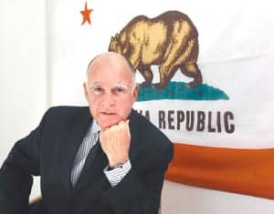 An older man in a dark suit and striped tie, possibly the Governor, is posing with his hand on his chin in front of a California state flag, which features a bear. CA Norml