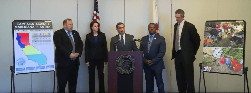 A group of five people stand behind a podium with microphones. To the left is a poster titled "Campaign Against Marijuana Planting" with maps and text, and to the right is an image of confiscated marijuana plants. Two flags are displayed in the background, as AG Becerra addresses the assembly. CA Norml