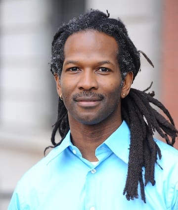 A person with long dreadlocks and a mustache, reminiscent of Drs. Carl Hart's style, is wearing a light blue button-up shirt. They are standing outdoors in front of a blurred building facade with a neutral expression and small hoop earrings in both ears. CA Norml