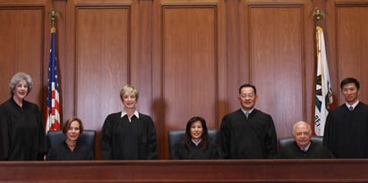 A group of seven judges, four standing and three seated, are posed in front of a wooden paneled background. The U.S. flag and a state flag are visible behind them. All judges, members of the Cal Supreme Court, are dressed in black judicial robes. CA Norml