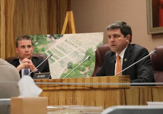 Two men in suits sit at a wooden panel during a state hearing. One is speaking into a microphone, discussing the drought's impact on local agriculture, while the other listens attentively. Behind them is a large, detailed photo of farmland or garden plots on an easel. CA Norml