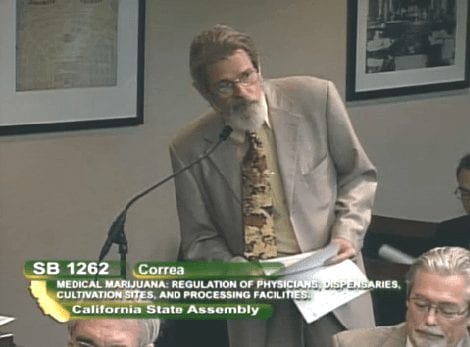 An individual in a suit speaks into a microphone at a podium with documents in hand. A legislative screen displays the medical marijuana regulation bill, SB 1262. Other seated individuals and framed pictures on the wall are visible in the background. CA Norml