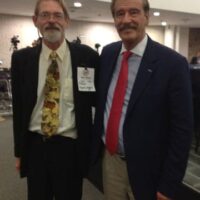 Two men are posing for a photo in a room at the US/Mexico Summit. One is wearing a light shirt, patterned tie, dark jacket, and name badge. The other man is in a dark jacket, light shirt, red tie, and light-colored pants. They are both smiling and standing side by side. CA Norml