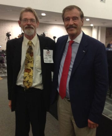 Two men are posing for a photo in a room at the US/Mexico Summit. One is wearing a light shirt, patterned tie, dark jacket, and name badge. The other man is in a dark jacket, light shirt, red tie, and light-colored pants. They are both smiling and standing side by side. CA Norml