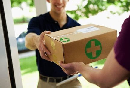 A delivery person from Cal NORML hands a brown package with a green medical cross symbol to a recipient at their doorstep. The delivery person is smiling, and the image is set outdoors with greenery visible in the background, highlighting the ease of home deliveries for cannabis products. CA Norml