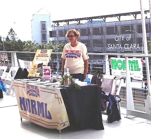 A person stands behind a table covered in a NORML tablecloth and various informational materials about California Law. They are wearing a t-shirt with a slogan. A sign reading "City of Santa Clara" is visible in the background along with the NORML banner. CA Norml