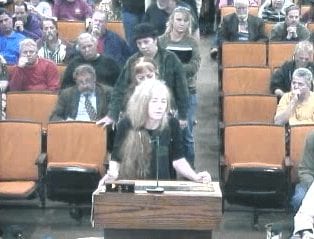 A person with long hair is speaking at a podium in a meeting room filled with attentive attendees. Two individuals stand behind the speaker, who seems to be discussing the "Tale of Two Counties". The audience appears engaged, seated in rows of orange chairs, perhaps in Fresno or El Dorado.
 CA Norml