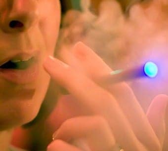 Close-up of a person holding a smokeless e-cig with a glowing blue tip, exhaling vapor. The background is blurred, emphasizing the person's mouth and the swirling vapor. CA Norml