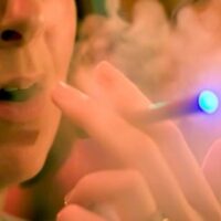 Close-up of a person holding and using an electronic cigarette. The device emits vapor that clouds the area around their face, with a blue light glowing from the tip. Their mouth is slightly open, suggesting an inhale or exhale of the vapor, potentially highlighting concerns addressed in CA Bill Against E-Cigarettes (SB 648). CA Norml