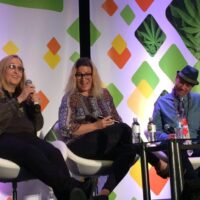 Three people are seated on stage in front of a colorful, geometric background with cannabis leaf designs at the NCIA conference. They are engaged in a panel discussion. The person on the left is speaking into a microphone, while the others listen attentively. Various drinks are on the table. CA Norml