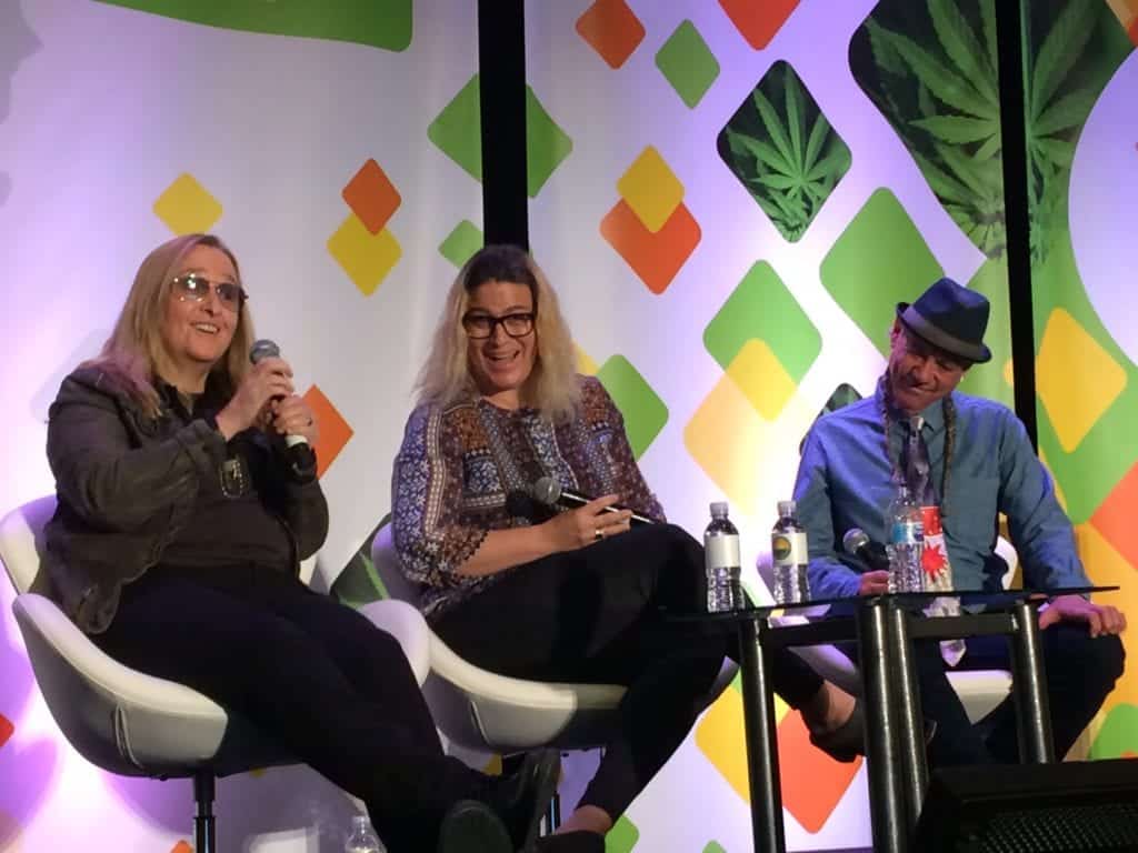 Three people are seated on stage in front of a colorful, geometric background with cannabis leaf designs at the NCIA conference. They are engaged in a panel discussion. The person on the left is speaking into a microphone, while the others listen attentively. Various drinks are on the table. CA Norml