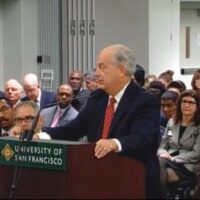 A man in a suit and red tie speaks at a podium labeled "University of San Francisco" in a crowded room. Many people are seated behind him, attentively listening as he discusses the recent Supreme Court ruling on the dispensary case. The room has a professional, formal setting. CA Norml