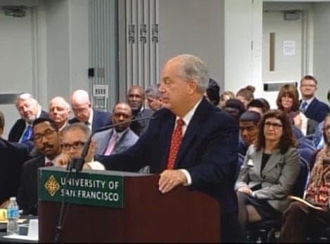 A man in a suit and red tie speaks at a podium labeled "University of San Francisco" in a crowded room. Many people are seated behind him, attentively listening as he discusses the recent Supreme Court ruling on the dispensary case. The room has a professional, formal setting. CA Norml