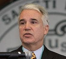 A man with short gray hair, wearing a dark suit, light blue shirt, and a yellow-and-black striped tie, speaks into a microphone. The background is blurred, showing part of a logo. He’s in San Francisco discussing efforts to erase minor marijuana convictions. CA Norml