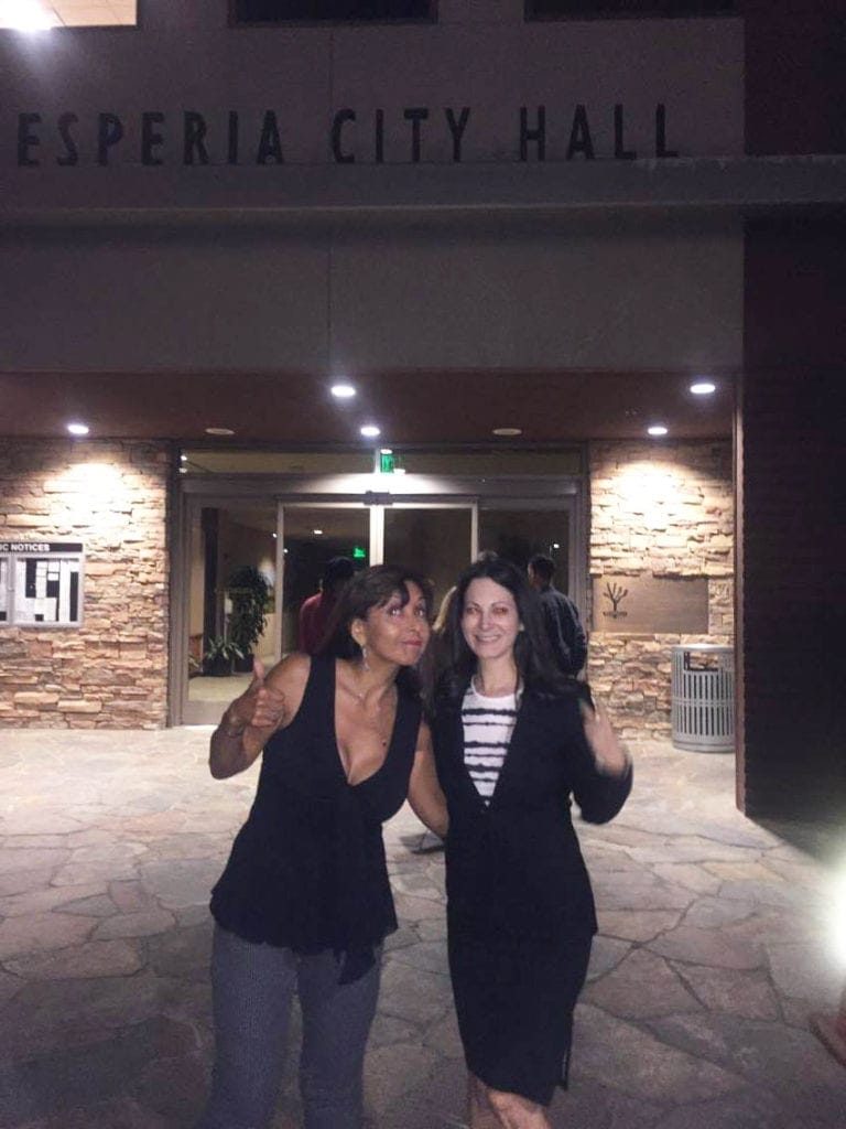 Two women are standing and posing with thumbs-up gestures outside a building at night. The building has a sign that reads "Hesperia City Hall" above the entrance, possibly discussing Hesperia Tables or Personal Cultivation. The scene is lit by exterior lights, highlighting the stone façade and glass doors behind them. CA Norml