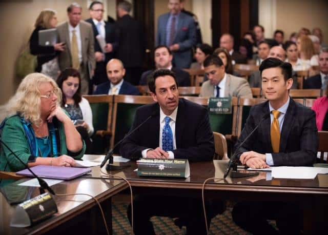 A group of people sit at a table in a busy room, likely a governmental or corporate meeting. Three individuals are in the foreground: a woman on the left, a man in the center speaking about the DUID Bill, and a man on the right. People in professional attire are seated and standing in the background. CA Norml