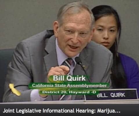 A man, identified by a name tag as Bill Quirk, California State Assemblymember for District 20, speaks at a Joint Legislative Committee Hearing about the Adult Use of Marijuana Act. Dressed in a gray suit and tie, he is seated in front of a microphone, with a woman visible behind him. CA Norml