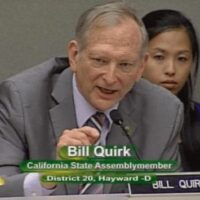 A man in a suit and tie, identified as Bill Quirk, is speaking at a desk with a microphone. A woman is seated behind him, attentively listening. The text on the screen reads, "Bill Quirk, California State Assemblymember, District 20, Hayward-D," during an event in Sacramento discussing Cannabis legislation. CA Norml