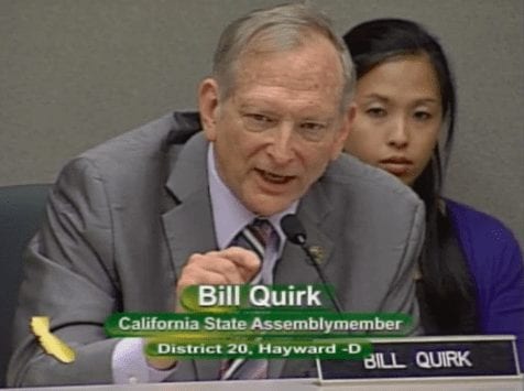 A man in a suit and tie, identified as Bill Quirk, is speaking at a desk with a microphone. A woman is seated behind him, attentively listening. The text on the screen reads, "Bill Quirk, California State Assemblymember, District 20, Hayward-D," during an event in Sacramento discussing Cannabis legislation. CA Norml