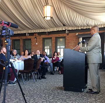 A man in a suit stands at a podium, speaking to an audience seated at round tables in a large, elegant room with draped ceilings. A camera on a tripod is set up in the foreground to record the event as he discusses reform in the California Legislature. The attendees are listening attentively. CA Norml