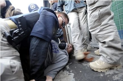 A law enforcement officer in tactical gear restrains a protestor on the ground during the Oaksterdam Protest. The restrained individual appears to have a partially bloodied face. Other officers and individuals in tactical gear are nearby. The setting appears to be outdoors near a building. CA Norml