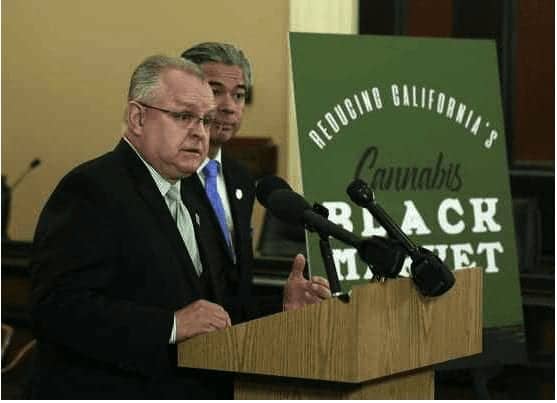 Two men at a podium during a press conference, standing in front of a sign that reads "Reducing California's Cannabis Black Market." The man on the left, wearing a suit and glasses, is speaking into multiple microphones about the new bill introduced to tackle cannabis taxes while the other man looks on. CA Norml