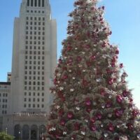 A large Christmas tree adorned with pink and silver ornaments stands prominently in front of a tall, white, art-deco style building under a clear blue sky. Recently featured by LA News, the tree is surrounded by colorful presents at its base. CA Norml