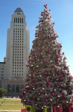 A large Christmas tree adorned with pink and silver ornaments stands prominently in front of a tall, white, art-deco style building under a clear blue sky. Recently featured by LA News, the tree is surrounded by colorful presents at its base. CA Norml